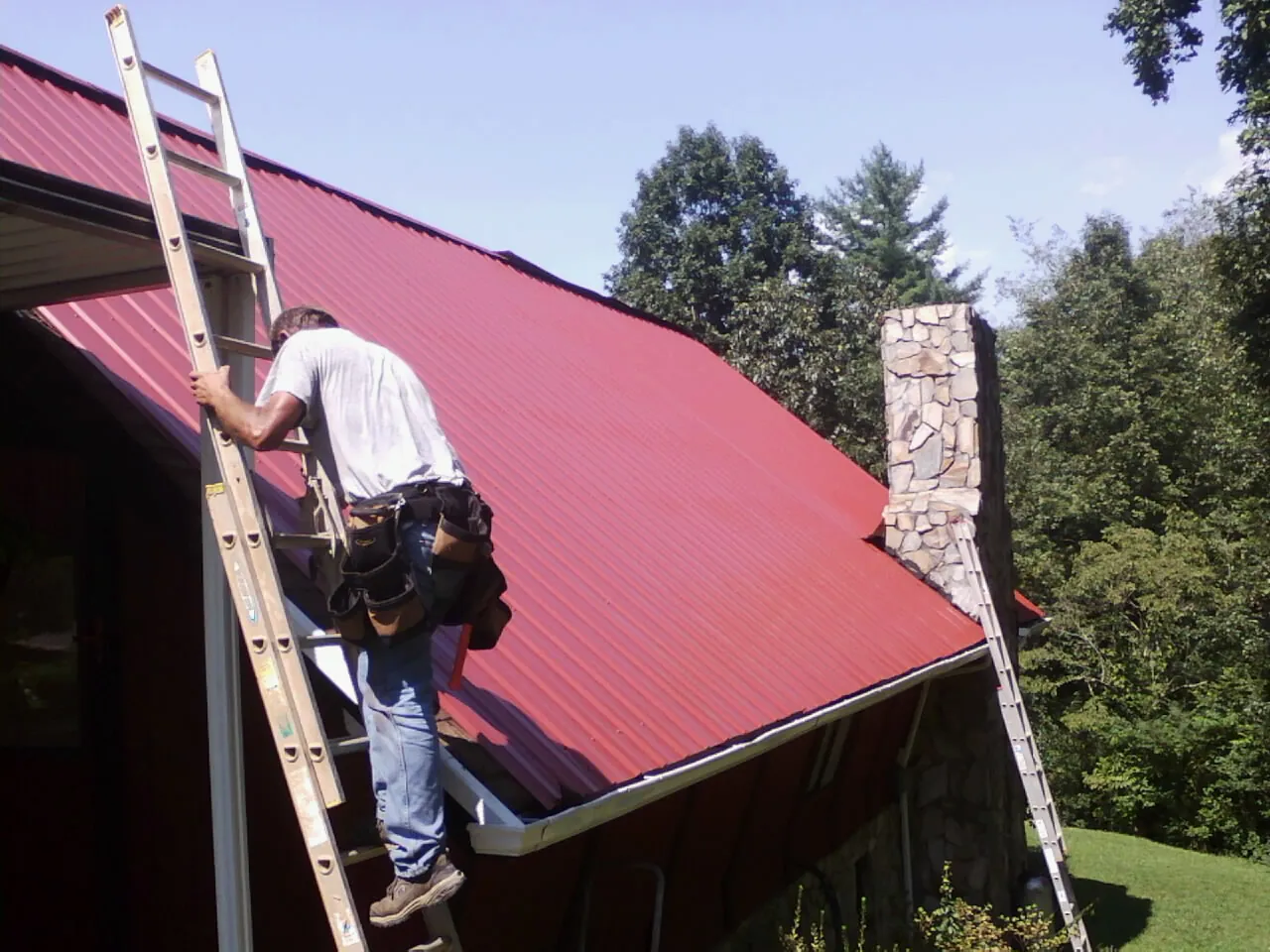 Roofer on a ladder beside a red metal roof and stone chimney — Soffit & Fascia Repair by Roofing Brothers in Parkton